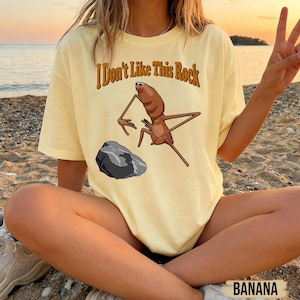 May include: A pale yellow short-sleeve t-shirt featuring a brown insect-like creature standing near a grey rock. The text "I Don't Like This Rock" is printed in brown letters above the graphic. The shirt has a crew neckline and a relaxed fit.