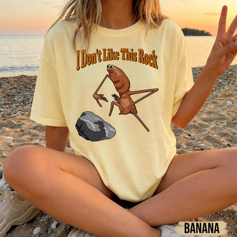 May include: A pale yellow short-sleeve t-shirt featuring a brown insect-like creature standing near a grey rock. The text "I Don't Like This Rock" is printed in brown letters above the graphic. The shirt has a crew neckline and a relaxed fit.