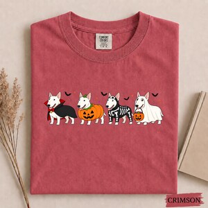 Puede incluir: Una camiseta de cuello redondo color carmes&iacute; con un gr&aacute;fico de cuatro perros Bull Terrier blancos disfrazados de Halloween: un vampiro, una calabaza, un esqueleto y un fantasma sosteniendo una calabaza, con peque&ntilde;os murci&eacute;lagos y estrellas.