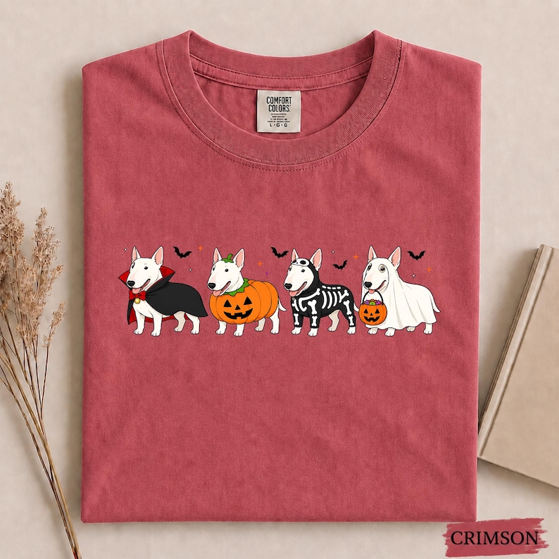 Puede incluir: Una camiseta de cuello redondo color carmes&iacute; con un gr&aacute;fico de cuatro perros Bull Terrier blancos disfrazados de Halloween: un vampiro, una calabaza, un esqueleto y un fantasma sosteniendo una calabaza, con peque&ntilde;os murci&eacute;lagos y estrellas.