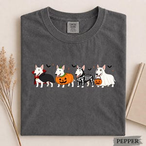 Puede incluir: Una camiseta gris carb&oacute;n con un gr&aacute;fico de cuatro perros Bull Terrier blancos con disfraces de Halloween. Los perros est&aacute;n vestidos de vampiro, calabaza, esqueleto y fantasma sosteniendo un cubo de calabaza, con peque&ntilde;os murci&eacute;lagos negros.