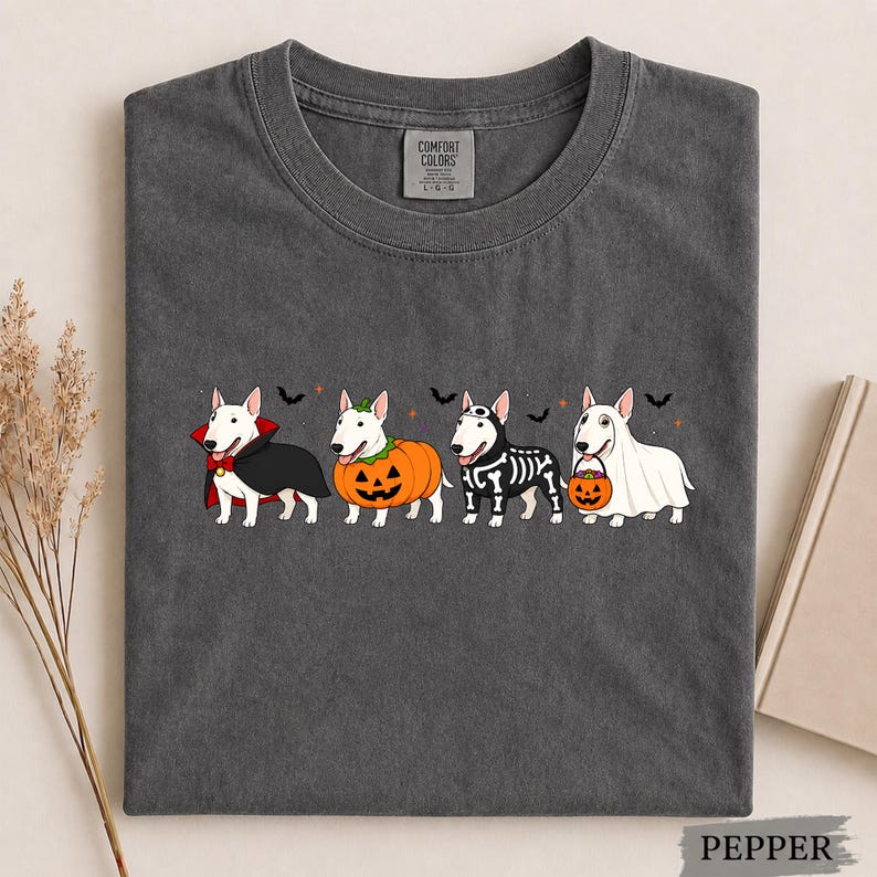 Puede incluir: Una camiseta gris carb&oacute;n con un gr&aacute;fico de cuatro perros Bull Terrier blancos con disfraces de Halloween. Los perros est&aacute;n vestidos de vampiro, calabaza, esqueleto y fantasma sosteniendo un cubo de calabaza, con peque&ntilde;os murci&eacute;lagos negros.