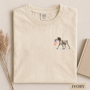 May include: An ivory-colored crew neck t-shirt featuring a small graphic on the left chest of a brown and white spotted dog carrying a small American flag in its mouth. The shirt has a Comfort Colors brand label printed on the inside of the collar.