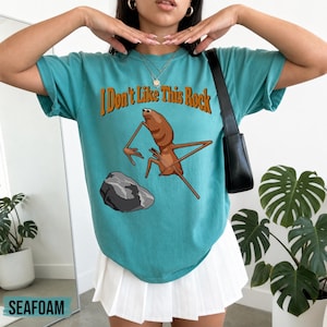 May include: A teal short-sleeve t-shirt featuring a graphic of a brown insect-like creature standing near a gray rock. The text "I Don't Like This Rock" is printed in orange above the creature. The shirt is paired with a white pleated skirt.