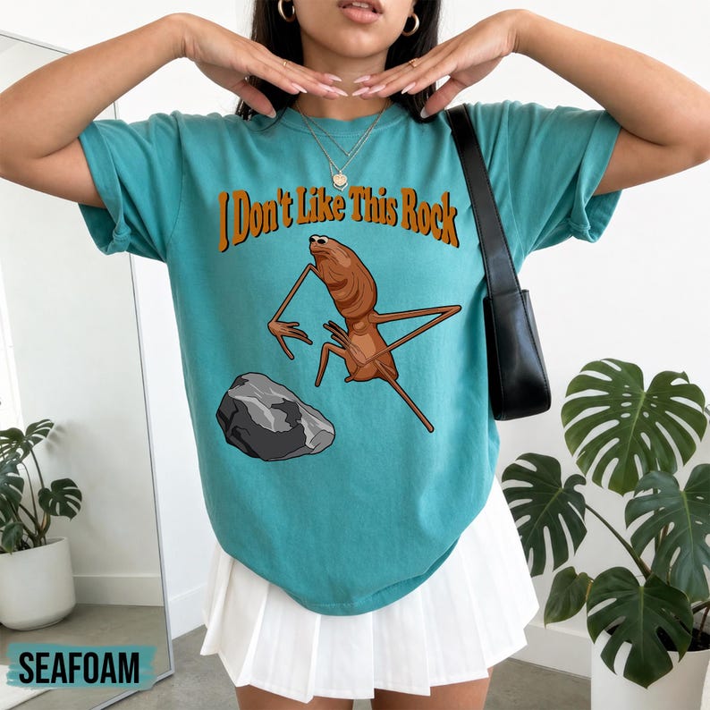 May include: A teal short-sleeve t-shirt featuring a graphic of a brown insect-like creature standing near a gray rock. The text "I Don't Like This Rock" is printed in orange above the creature. The shirt is paired with a white pleated skirt.