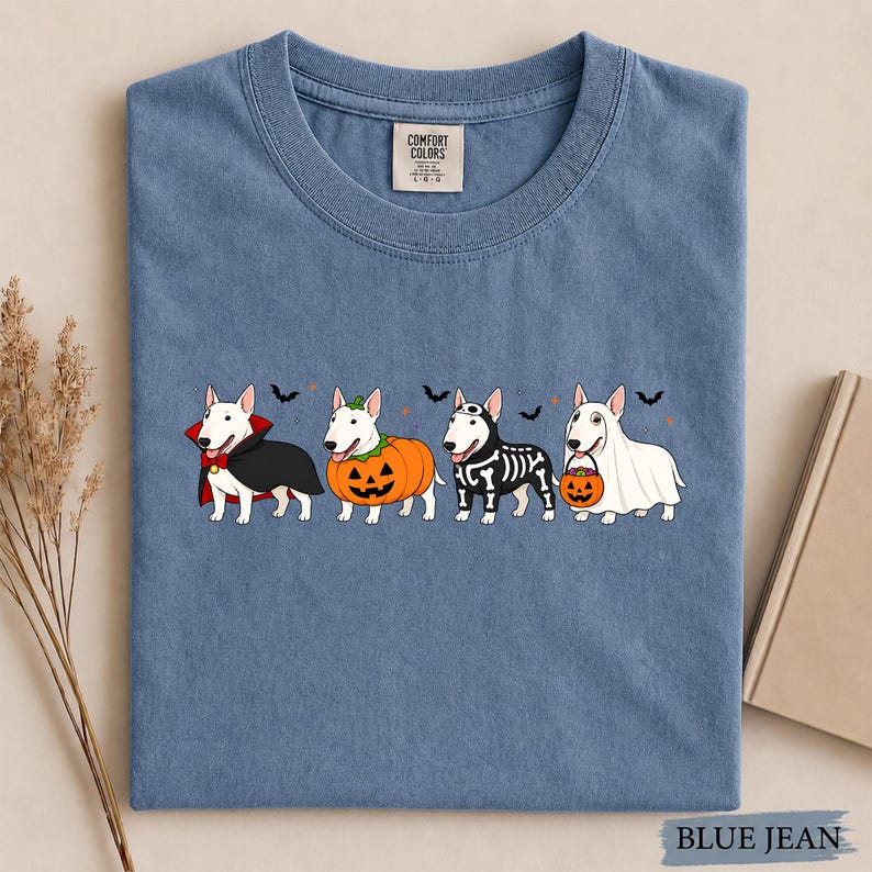 Puede incluir: Una camiseta azul de cuello redondo con un gr&aacute;fico de cuatro perros bull terrier blancos con disfraces de Halloween, incluyendo un vampiro, una calabaza, un esqueleto y un fantasma, con peque&ntilde;os murci&eacute;lagos negros y puntos naranjas.