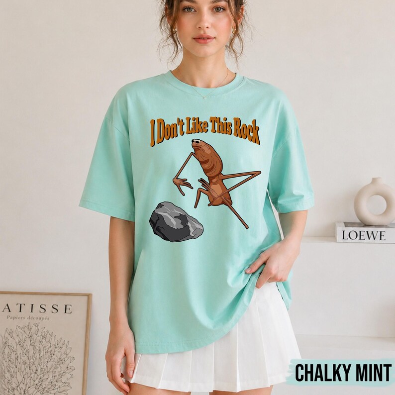 May include: A light mint green short-sleeve t-shirt featuring a brown insect illustration and a grey rock graphic. The text "I Don't Like This Rock" is printed in brown above the insect. The shirt has a crew neckline and a relaxed fit.