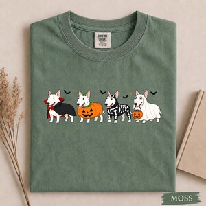 Puede incluir: Una camiseta verde musgo con un gr&aacute;fico de cuatro perros bull terrier blancos con disfraces de Halloween. Los perros est&aacute;n vestidos de vampiro, calabaza, esqueleto y fantasma, acompa&ntilde;ados de peque&ntilde;os murci&eacute;lagos negros y estrellas naranjas.