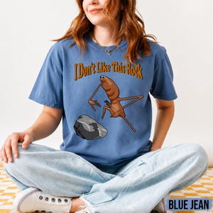 May include: A blue short-sleeved t-shirt featuring a graphic of a brown insect-like creature standing near a grey rock. Above the creature, the text reads "I Don't Like This Rock" in a brown, stylized font. The shirt has a crew neckline.