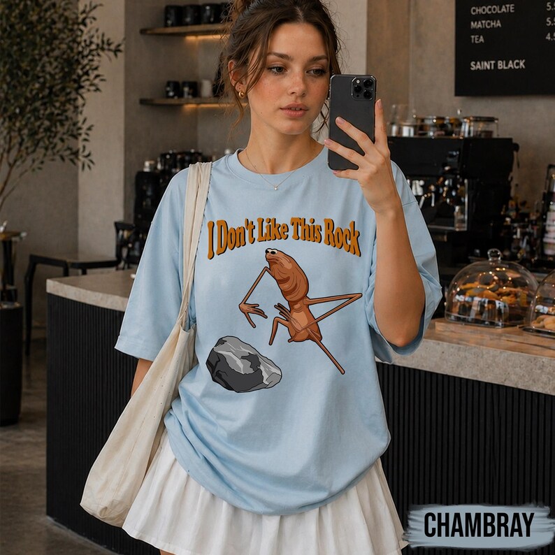 May include: A light blue t-shirt featuring a graphic of a brown, long-limbed creature standing near a grey rock. The text "I Don't Like This Rock" is printed in orange above the creature. The shirt has short sleeves and a crew neckline.