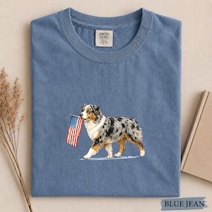 May include: A blue jean colored t-shirt with a Comfort Colors label at the neck. The center graphic features a merle Australian Shepherd dog walking while holding a small American flag in its mouth. The dog has white, black, and tan fur.