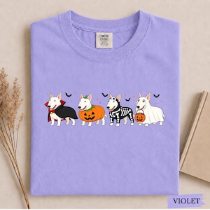 Puede incluir: Una camiseta de color violeta claro con un gr&aacute;fico de cuatro perros Bull Terrier blancos vestidos con disfraces de Halloween: un vampiro, una calabaza, un esqueleto y un fantasma. La camiseta tiene cuello redondo y etiqueta Comfort Colors.