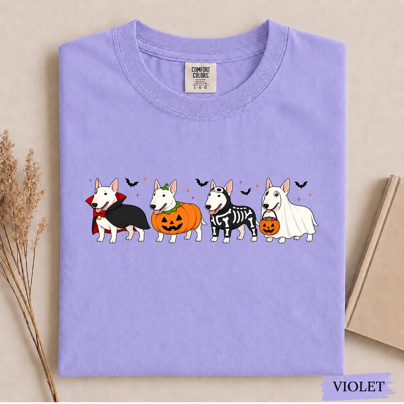 Puede incluir: Una camiseta de color violeta claro con un gr&aacute;fico de cuatro perros Bull Terrier blancos vestidos con disfraces de Halloween: un vampiro, una calabaza, un esqueleto y un fantasma. La camiseta tiene cuello redondo y etiqueta Comfort Colors.