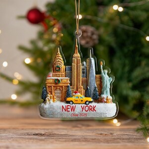 Puede incluir: Adorno navideño de acrílico transparente con el horizonte de Nueva York, la Estatua de la Libertad y un taxi amarillo. Se ve el texto "NEW YORK Chloe 2025". El adorno cuelga frente a un árbol de Navidad.