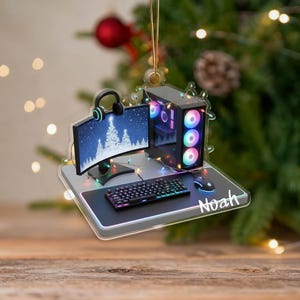 May include: A clear acrylic Christmas ornament depicting a gaming setup. The ornament includes a computer monitor, keyboard, mouse, and PC tower with colourful lights. The name "Noah" is written on the mousepad. A headset is also included.