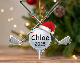 Puede incluir: Un adorno navideño de acrílico transparente con una pelota de golf con un gorro de Papá Noel, palos de golf cruzados y luces de cadena. La pelota de golf tiene el nombre "Chloe" y el año "2025" impresos. El adorno cuelga de una cuerda dorada.