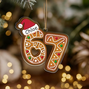 Könnte beinhalten: Ein weihnachtlicher Christbaumschmuck in Form der Zahl 67, gestaltet wie ein Lebkuchen. Mit dem Namen "Chloe" und dem Jahr "2025" auf einer Weihnachtsmütze, verziert mit roten und grünen Details.
