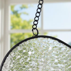 May include: A circular suncatcher featuring a textured, translucent glass panel with a pebbled, light green and clear appearance. The glass is encased in a thin black metal frame and is suspended by a matching black metal chain link.