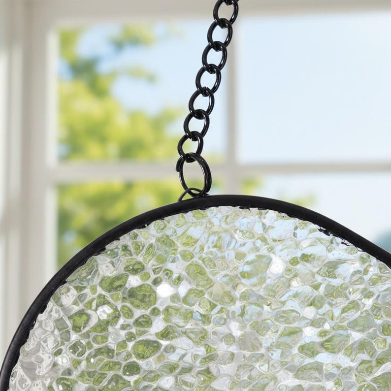 May include: A circular suncatcher featuring a textured, translucent glass panel with a pebbled, light green and clear appearance. The glass is encased in a thin black metal frame and is suspended by a matching black metal chain link.