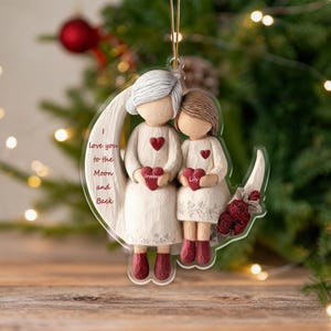 Może przedstawiać: Przezroczysta ozdoba akrylowa przedstawiająca dwie postacie, jedną z siwymi włosami, a drugą z brązowymi, trzymające przedmioty w kształcie serca. Ozdoba głosi "I love you to the Moon and Back" i zawiera imiona "Grandma" i "Lily".