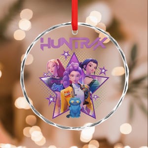 May include: A clear glass ornament with a red ribbon. The ornament features a graphic of three animated women inside a purple star, with the word "HUNTRAX" above them. A blue cat is in the foreground.