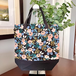 May include: A tote bag featuring a black background with a floral pattern of pink roses, blue leaves, and yellow berries. The bag has a solid black bottom panel and two black fabric handles attached to the top. The design is consistent throughout.