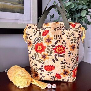 Peut inclure: Un sac à cordon en tissu avec un motif floral composé de fleurs rouges, orange et brunes sur un fond jaune clair. Le sac possède des poignées en tissu vert olive et des cordons de serrage jaunes. Une pelote de laine jaune est posée devant.