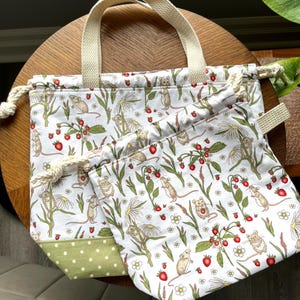 May include: A white fabric tote bag and a matching drawstring pouch featuring a repeating pattern of small brown mice, red strawberries, and white daisy flowers. The tote bag has cream-coloured woven handles and a light green polka dot base panel.