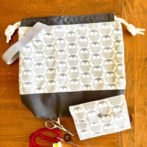 May include: A grey drawstring bag with a penguin pattern and a matching grey pouch. The bag has a grey strap and white drawstrings. The pouch has a button closure. There are also red yarn, scissors, and a spool of thread on a wooden surface.
