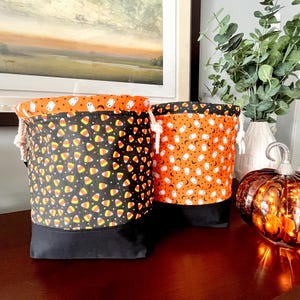 May include: Two fabric drawstring bags featuring Halloween-themed patterns. The left bag has a black base with a candy corn print and an orange top with white ghosts. The right bag has a black base with an orange top featuring white ghosts.