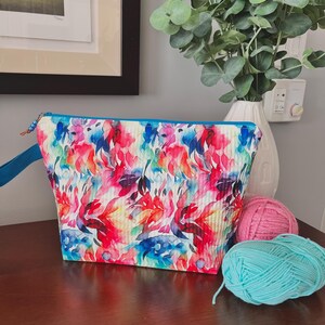 May include: A trapezoidal zipped pouch featuring a vibrant watercolour-style floral pattern in shades of pink, red, blue, and purple on a white background. The bag includes a blue fabric wrist strap and a blue zip closure with a small bead accent.