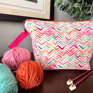 May include: A quilted zip pouch featuring a multi-coloured chevron pattern in pink, orange, blue, and green. The pouch has a bright pink wrist strap with colourful beads, accompanied by three balls of yarn and two dark pink knitting needles.
