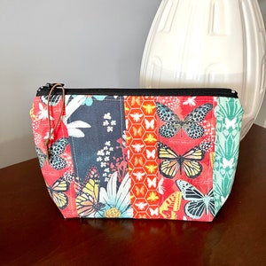 Peut inclure: Une petite pochette rectangulaire zippée avec un motif patchwork représentant des papillons, des abeilles et des fleurs. La pochette est en tissu coloré avec une fermeture éclair noire et une tirette en cuir marron.
