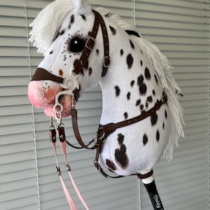 May include: A white hobby horse with black spots, a white yarn mane, and a brown leather bridle with a white bit. The horse has a pink-tinted muzzle and is attached to a black stick featuring the white text Hobby Horse with a small horse icon.