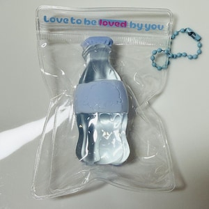 May include: A clear, bottle-shaped keyring with a pale blue cap and band, inside a transparent plastic pouch. The pouch has the text "Love to be loved by you" printed on it. A pale blue beaded chain is attached.