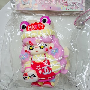 May include: A colorful, cartoon-style figurine of a girl with pink hair, wearing a red dress and holding a Hello Kitty bag. The figurine has a frog-like hat with the word "HAPPY" on it. The figurine is packaged in a clear plastic bag.