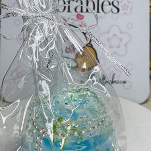 May include: A blue decorative jar with a fish tank design, embellished with rhinestones and pearls, wrapped in clear cellophane and tied with a silver ribbon. The background features a white card with the text "Affordable Memorables".