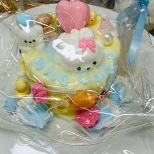 May include: A small, decorated cake wrapped in clear cellophane. The cake is yellow with blue and white frosting, featuring a cartoon bunny and the words "Nice Cake". It is adorned with pink hearts, bears, and other colorful decorations. A blue ribbon is tied to the cellophane.