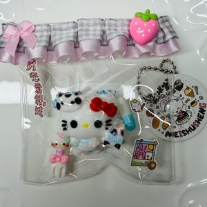 May include: A clear plastic bag containing a white Hello Kitty figure with a red bow, a pink cat, and a blue milk bottle. The bag is decorated with a pink bow and a pink strawberry charm. A small tag with food illustrations hangs from a chain.