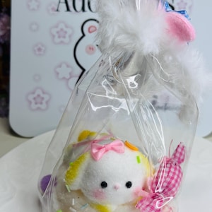 Può includere: Un sacchetto di cellophane trasparente legato con finta pelliccia bianca e un fiocco rosa, contenente un coniglietto di peluche con un fiocco rosa, zuccherini e una caramella rosa e bianca a quadretti. Lo sfondo ha il testo "Affordable Adorable".