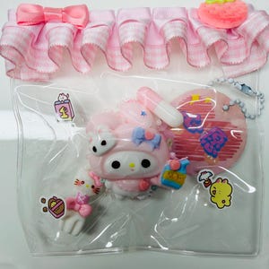May include: A clear plastic bag containing several pink and white My Melody themed accessories. The accessories include a pink ruffle trim, a My Melody figure, a Hello Kitty figure, and other small charms. The bag is sealed with a pink ribbon.