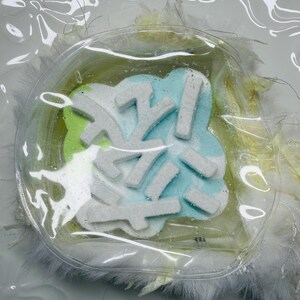 May include: A decorative item featuring a snowflake design. The snowflake is white and sits on a light blue and green background, encased in clear plastic. The item is surrounded by white feathers and rests on a white surface.