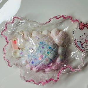 Pode incluir: Um item em forma de caranguejo em tons pastel com glitter e confetes em forma de estrela, embalado em um saco plástico transparente com uma borda rosa. O item tem um adesivo Hello Kitty.