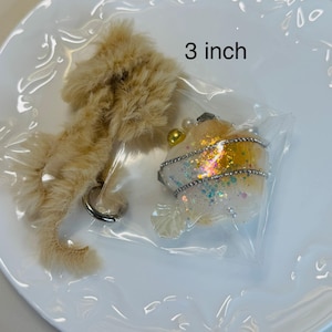 May include: A beige, fluffy keychain and a decorative resin item, both on a white plate. The resin item, approximately 3 inches in size, is encased in plastic and features glitter and embellishments. The keychain has a silver ring.