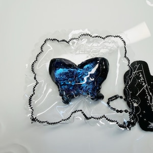 May include: A black and blue butterfly-shaped pendant with a glittery, galaxy-like design. The pendant is encased in a clear, scalloped-edged plastic. A black beaded chain and a black tag with white text are attached.
