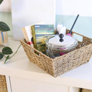 Può includere: Un cesto rettangolare intrecciato pieno di libri, una pentola di vetro con coperchio e candele. Il cesto è realizzato in materiale intrecciato color naturale. La pentola di vetro ha un coperchio argentato con un pomello nero. È visibile un utensile nero per mescolare.