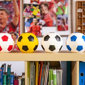 Op de afbeelding: Vier voetballen op een rij, elk met een ander kleurenschema: rood en wit, geel en zwart, wit en zwart en blauw en wit. Ze zijn gerangschikt op een lichtgekleurde houten plank, met boeken en mappen eronder.