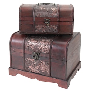 Può includere: Due forzieri in legno marrone scuro, uno impilato sull'altro. Presentano pannelli metallici floreali decorativi e chiusure in metallo. Il forziere superiore ha una maniglia. I forzieri hanno un aspetto vintage e antico.
