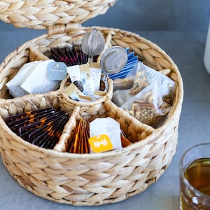 Può includere: Un organizer per tè rotondo in giacinto d'acqua intrecciato con coperchio incernierato, dotato di molteplici scomparti interni riempiti con varie bustine di tè, bustine di zucchero e due infusori a rete in acciaio inossidabile al centro.