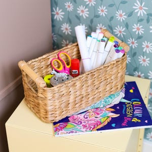 Può includere: Un cestino rettangolare in giunco intrecciato con due manici in legno chiaro contenente vari materiali artistici, tra cui pennarelli bianchi, una colla stick Pritt rossa, forbici rosa e una piccola statuetta di zebra, accanto a un libro.
